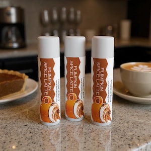 May include: Three white lip balm tubes with labels that read "Pumpkin Spice Latte." The labels feature a graphic of a latte and a brown and white background. A slice of pie and a cup of coffee are in the background.