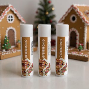 May include: Three white lip balm tubes with brown labels that read "GINGERBREAD" and feature a gingerbread house illustration. The background includes gingerbread houses and a small Christmas tree, creating a festive holiday theme.