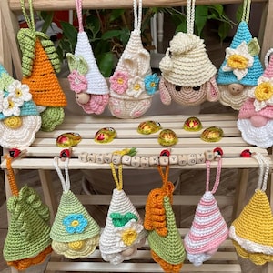 Może przedstawiać: Kolekcja 12 szydełkowych krasnoludków, każdy z innym wzorem i kolorystyką. Krasnoludki wiszą na drewnianej półce i są ozdobione kwiatami, liśćmi i innymi dodatkami. Krasnoludki mają wszystkie czapki i długie brody.