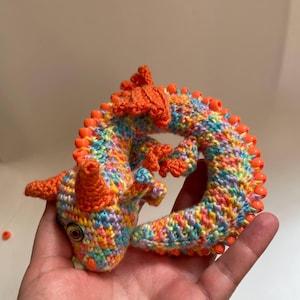 A fully crocheted sensory dragon curled into a coil resting in an open adult palm, demonstrating its pocket-sized, compact form.