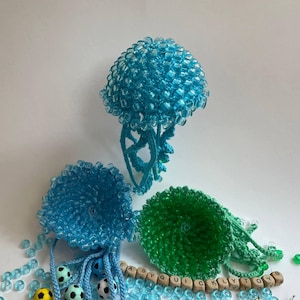Op de afbeelding: Drie kwalvormige ornamenten gemaakt van blauwe en groene kralen en draad. Twee zijn lichtblauw en één is groen, elk met een afgeronde bovenkant en bungelende tentakels. Een snoer van houten letterkralen met tekst is inbegrepen.