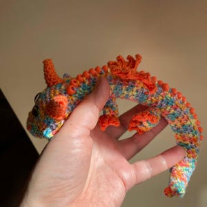 A hand holding a multicolor crochet dragon with orange beaded wings, stretched out straight between fingers to show the full length of its flexible, articulated body.