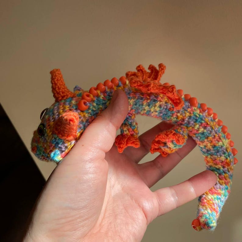 A hand holding a multicolor crochet dragon with orange beaded wings, stretched out straight between fingers to show the full length of its flexible, articulated body.