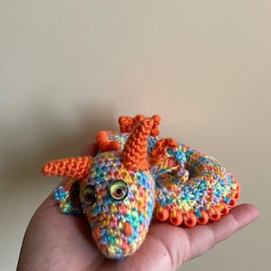 Front view of a multicolor dragon with orange beaded wings resting in a curled ball on an open palm after play, emphasizing its pocket-sized, coiled form.