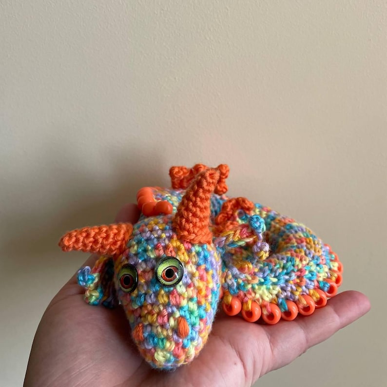 Front view of a multicolor dragon with orange beaded wings resting in a curled ball on an open palm after play, emphasizing its pocket-sized, coiled form.