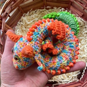 A different angle of the multicolor dragon with orange beaded wings curled in a palm, with a wicker basket softly visible in the background, highlighting its small size and texture.