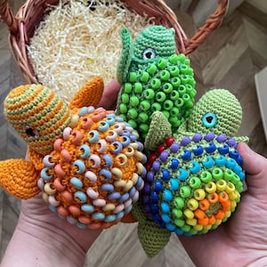 May include: Three handmade crochet turtle toys in vibrant colors. One is orange with pastel beads, another is green with green beads, and the third is green with rainbow beads. The turtles are held in a person's hands.