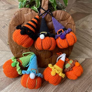 May include: Seven crocheted pumpkin ornaments with different coloured hats. The pumpkins are orange and the hats are green, blue, purple, black, and striped. The ornaments are sitting on a wooden surface.