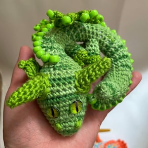 A green crochet sensory dragon curled into a coil resting in an open palm, viewed from the front and slightly above, set against a clean neutral background.