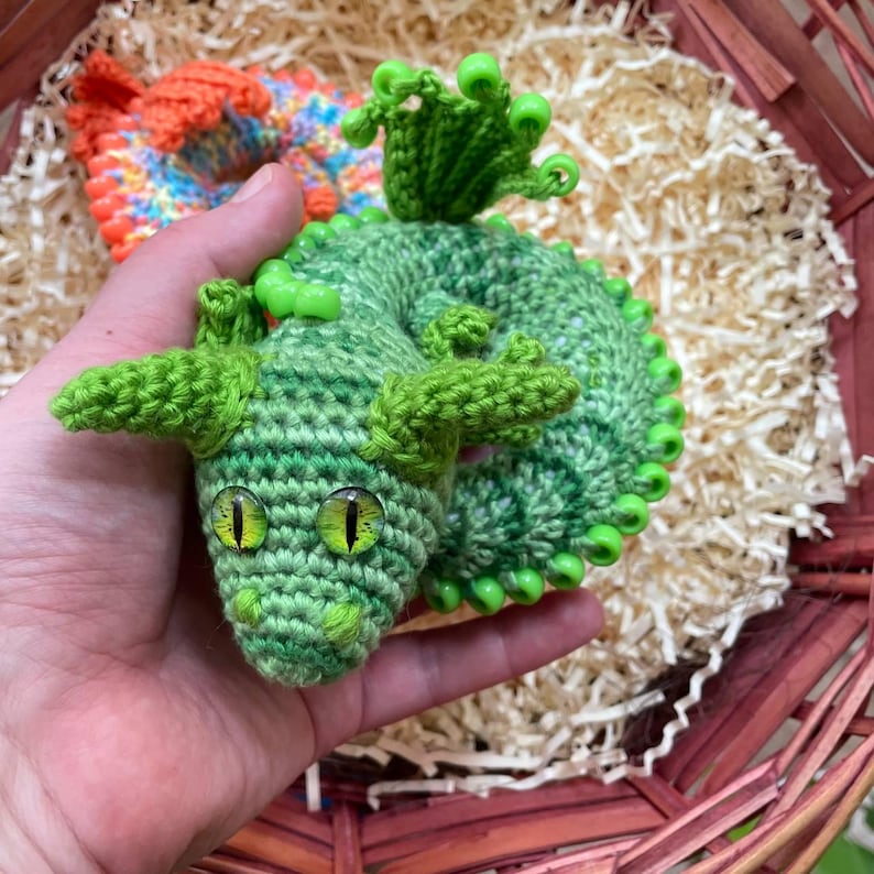 A green sensory dragon coiled in a hand, photographed from the front with a wicker basket softly visible in the background, highlighting its compact size.