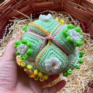 Puede incluir: Una bola hecha a mano, tejida a ganchillo en tonos de verde, amarillo, rosa y blanco. La bola presenta un diseño geométrico con acentos florales y adornos de cuentas. El proyecto artesanal se sostiene en una mano.