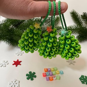 May include: Three green beaded Christmas tree decorations with colourful star accents hanging from green cords. The decorations are arranged in a row and are held by a hand. The words "EASY QUICKLY TOYS" are spelled out in colourful letter tiles below the decorations.