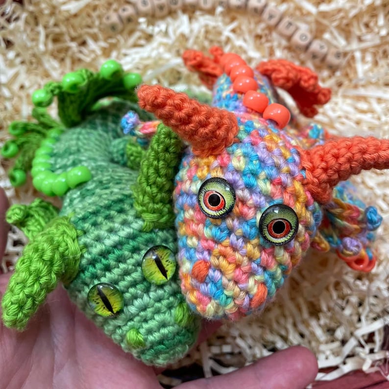 Two crochet dragons (multicolor and green) held together in a single palm, viewed from the front and slightly above, with a wicker basket in the background.