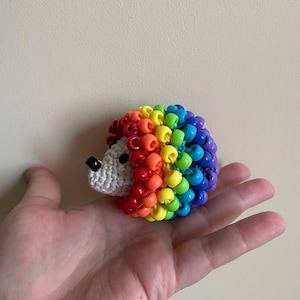 May include: A colourful hedgehog ornament made with beads. The body is covered in rainbow-coloured beads, transitioning from red to purple. The face is white with a black nose. A unique decorative item, perfect for a child's room or as a quirky gift.