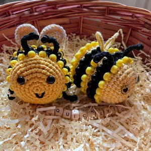 Peut inclure: Deux abeilles faites au crochet, jaunes et noires, avec des détails de perles noires, dans un panier. L'une des abeilles a un visage souriant. Elles sont posées sur des copeaux de papier. Des lettres en bois épellent "QUICKLY TO".