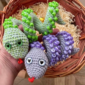 May include: Handmade crochet snakes in a basket. The snakes are in shades of green, gray, and purple, with heart-shaped eyes and red tongues. The snakes are decorated with small beads. The basket is made of woven wood.