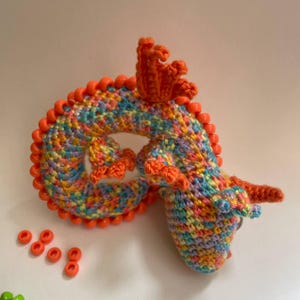 A multicolor crochet sensory dragon coiled into a circle on a neutral background, with a few loose pony beads placed next to it, showing its compact size.