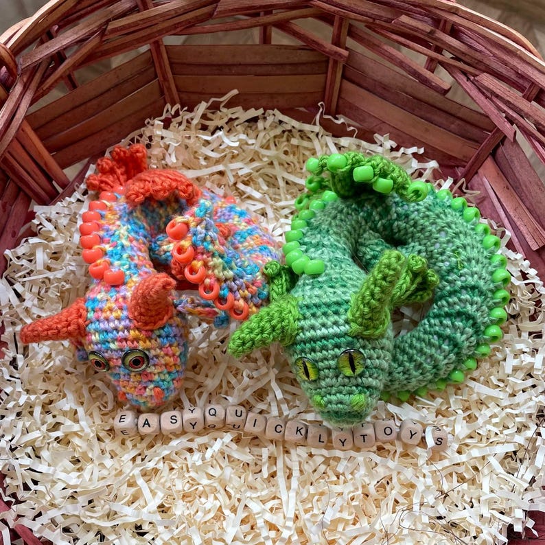 Two finished crochet dragons (orange and green) nestled together in a small wicker basket, presented as a handmade gift idea.