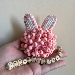 May include: A handmade pink beaded bunny with white and pink crochet ears. The bunny is surrounded by the words "EASY QUICKLY" spelled out on small wooden blocks. The bunny is a unique craft item.