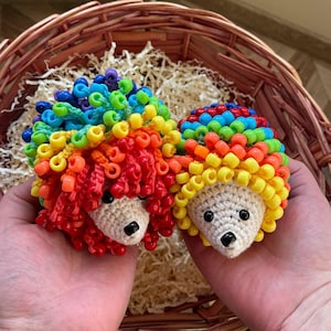 Puede incluir: Dos erizos de ganchillo hechos a mano con púas de cuentas de colores arcoíris. Los erizos tienen ojos de cuentas negras y un hocico beige. Se sostienen frente a una cesta de mimbre llena de papel triturado.