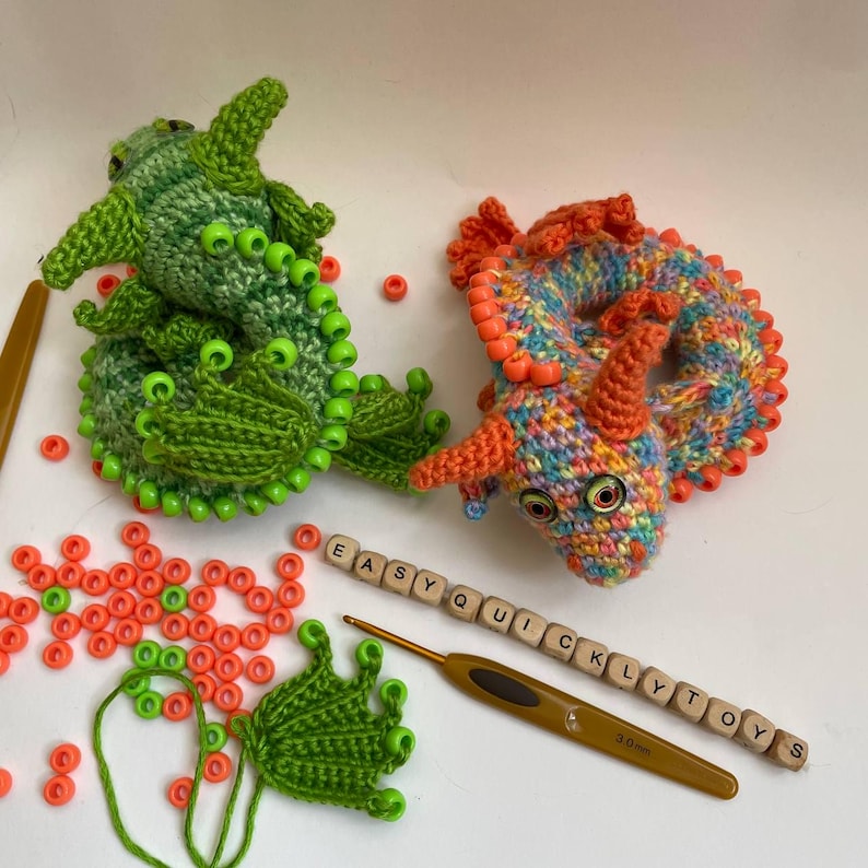 Two finished crochet sensory dragons side by side on a neutral background. The left dragon has orange beads and orange wings, the right dragon is green with lighter green wings. A crochet hook, pony beads pile, and a detached beaded wing are placed nearby.