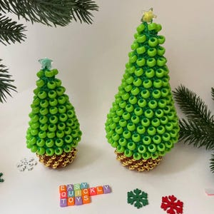 May include: Two handmade Christmas tree ornaments. The trees are green with a gold base and star toppers. The word "EASY QUICKLY TOYS" is spelled out with colorful letter blocks. Decorative snowflakes and pine branches are also visible.