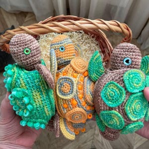 May include: Three handmade crochet turtle toys in shades of green, brown, and orange. Each turtle has a detailed shell and head with button eyes. The turtles are displayed in a woven basket, showcasing the craftsmanship of the crochet work.