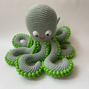 May include: A gray crocheted octopus with green beads around each tentacle. The octopus has white eyes and a red smile.