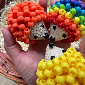 May include: A collection of handmade beaded hedgehog ornaments in various colors. The hedgehogs have crocheted bodies and bead quills in orange, rainbow, and yellow. Each has black bead eyes and nose.