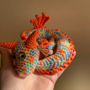 May include: A colorful, crocheted dragon figurine with orange horns, wings, and accents. The dragon's body is a mix of rainbow colors, and it has large, round eyes. The dragon is curled up, and the details are clearly visible.