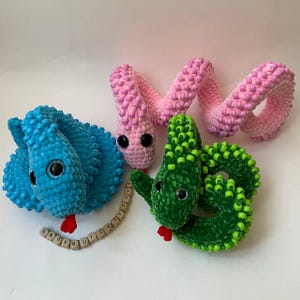 Puede incluir: Tres juguetes de serpiente de crochet en rosa, azul y verde. Cada serpiente tiene una textura de cuentas y grandes ojos negros. La serpiente azul tiene las palabras "EASY QUICK TOYS" deletreadas en bloques de madera.