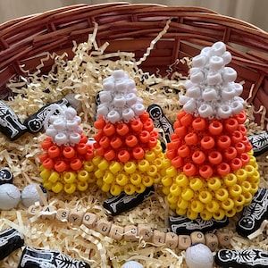 Puede incluir: Tres decoraciones de caramelos de maíz hechas a mano en una cesta tejida llena de papel triturado. El caramelo de maíz es amarillo, naranja y blanco. También se incluyen pequeñas decoraciones con temas de esqueletos y globos oculares.