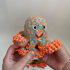 Puede incluir: Un juguete de pulpo tejido a ganchillo con hilo multicolor en tonos rosa, azul, amarillo y naranja. El pulpo presenta dos grandes ojos de plástico con pupilas azules y una boca roja bordada. Cada tentáculo está adornado con cuentas naranjas.