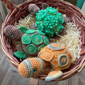 May include: A collection of handmade crochet turtle toys in a woven basket. The turtles feature various colors, including brown, green, orange, and blue. The basket is filled with paper filler, creating a cozy display. The turtles are a charming, handcrafted item.