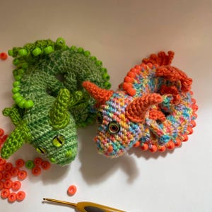 Two crochet dragons (multicolor and green) placed side by side in coiled positions, with a pile of pony beads and a crochet hook lying next to them on a flat surface.