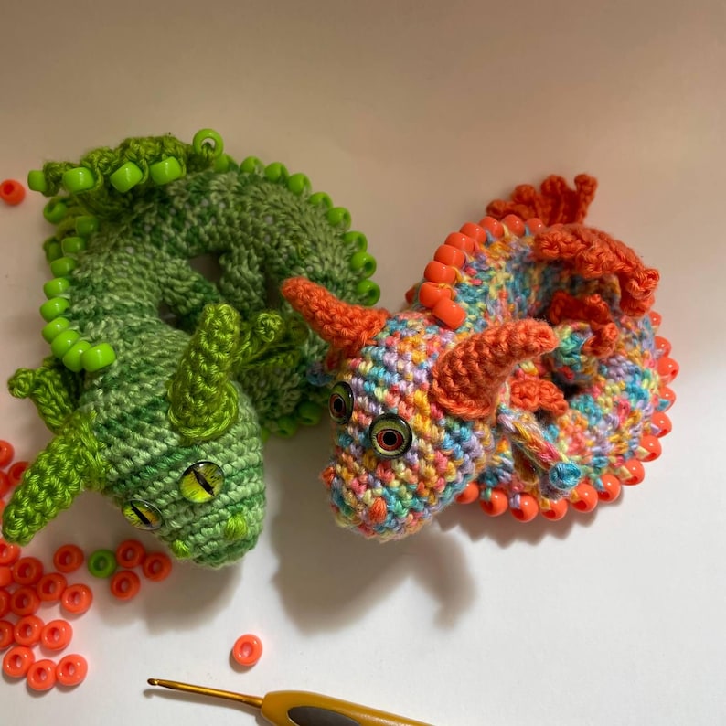 Two crochet dragons (multicolor and green) placed side by side in coiled positions, with a pile of pony beads and a crochet hook lying next to them on a flat surface.