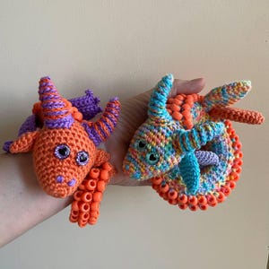 May include: Two crocheted dragon figures featuring yarn and plastic beads. One dragon is orange with purple horns and heart-shaped eyes. The second dragon is multicolored with blue horns and heart-shaped eyes, accented with orange plastic beads.