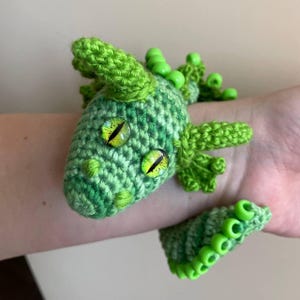 Side view of a green crochet dragon with beaded crest and lighter green wings, its body slightly curled, resting on a neutral background.