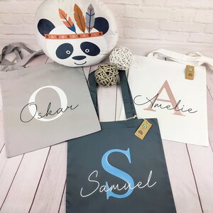 Könnte beinhalten: Drei Tragetaschen mit personalisierten Namen und Anfangsbuchstaben. Die Taschen sind aus natürlichem Canvas-Gewebe und haben eine hellbraune Farbe. Die Namen sind "Oskar", "Amelie" und "Samuel".