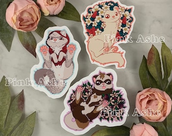 May include: Three illustrated stickers featuring ferrets in different poses. The first sticker shows a ferret wearing a red hooded sweater. The second sticker shows a ferret with white fur sitting in a bed of pink and white flowers. The third sticker shows a brown ferret with a pink flower crown.