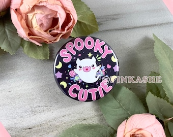 May include: A black and pink button with a white ghost and the text "Spooky Cutie" in pink letters. The ghost has pink cheeks and is surrounded by stars, hearts, and candy.