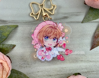May include: A pink and white anime character charm with a gold bow keychain. The character is holding a pink and white stuffed animal and has a pink hat with a white bow. The charm has a sparkly background with pink flowers and a gold star.