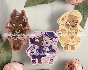 May include: Three sticker designs featuring cute cartoon characters in various poses. The first sticker features a brown demon with horns and a pink dress. The second sticker features a pair of purple and black demons with wings and black dresses. The third sticker features a yellow and white demon with wings and a yellow dress.