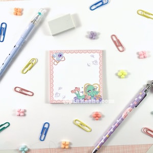 May include: A pink notepad with a white border and a cute cartoon illustration of a green octopus with pink hair and a gold crown. The octopus is wearing a pink dress and holding a pink heart. The notepad is on a white surface with colorful paper clips and flowers.