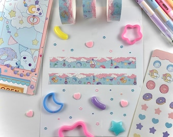 May include: A flat lay of pastel blue and pink washi tape, star and moon shaped cookie cutters, and pastel colored stickers with a cloud and moon design. The washi tape has a repeating pattern of clouds, stars, and moons.