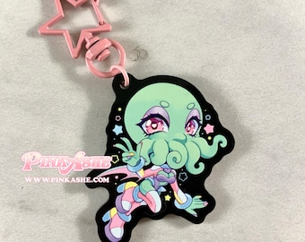 May include: A black keychain featuring a cartoon octopus-like character with a light green head, pink eyes, and colorful clothing. The keychain has a pink star-shaped clasp and the text "PINKASHE" and "WWW.PINKASHE.COM".
