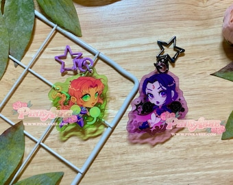 May include: Two acrylic keychains featuring chibi-style characters. One keychain has a purple star clasp and a character with long red hair on a green background. The other has a black star clasp and a character with purple hair on a pink background.