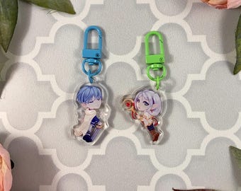May include: Two character charms with blue and green metal keychains. The charms are clear acrylic with printed images of anime characters. One character is a blue-haired boy wearing a white robe and the other is a girl with long white hair wearing a white dress and holding a golden staff.