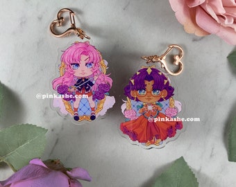 May include: Two colorful anime-style character charms with gold keychains. The charm on the left features a pink-haired character with a sword, wearing a blue and gold outfit. The charm on the right features a purple-haired character with a red and gold outfit, holding a glowing orb. Both charms have a white background with pink roses and the text "@pinkashe.com".