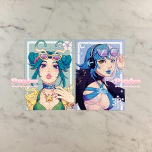 May include: Two art prints featuring stylized female characters with blue and green hair, sunglasses, and fantasy-themed attire. The prints have a light blue border and the text "Pinky Ashe" and "www.pinkashe.com".